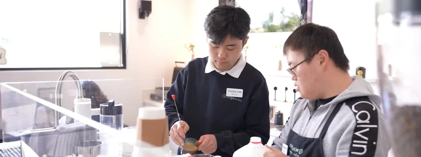 Two young men in Seesaw aprons preparing coffee together at a cafe counter.